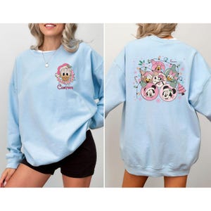 May include: Light blue sweatshirt featuring a cartoon character in a Santa hat and the word "Custom" on the front. The back of the sweatshirt displays a festive design with Mickey and Minnie Mouse, Daisy Duck, and other characters in Christmas ornaments.