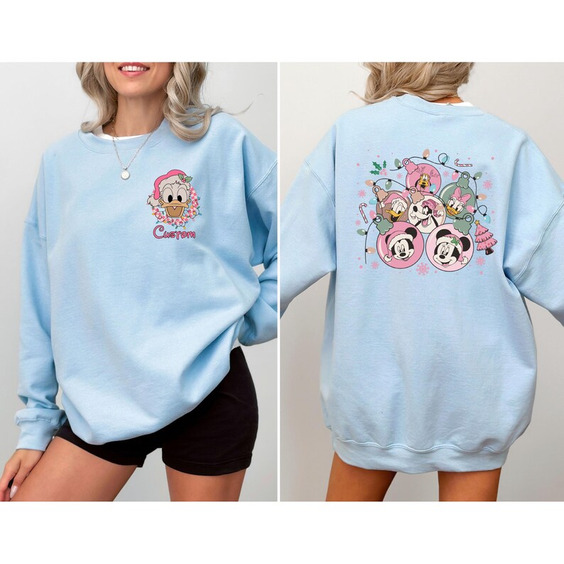 May include: Light blue sweatshirt featuring a cartoon character in a Santa hat and the word "Custom" on the front. The back of the sweatshirt displays a festive design with Mickey and Minnie Mouse, Daisy Duck, and other characters in Christmas ornaments.