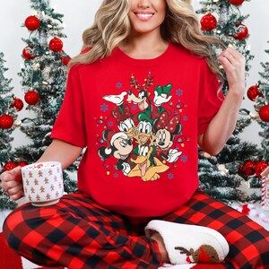 May include: A red t-shirt featuring a festive design with Mickey Mouse, Minnie Mouse, Donald Duck, Daisy Duck, Goofy, and Pluto. The shirt is paired with red and black plaid pajama pants. A white mug with gingerbread men is held in the hand.