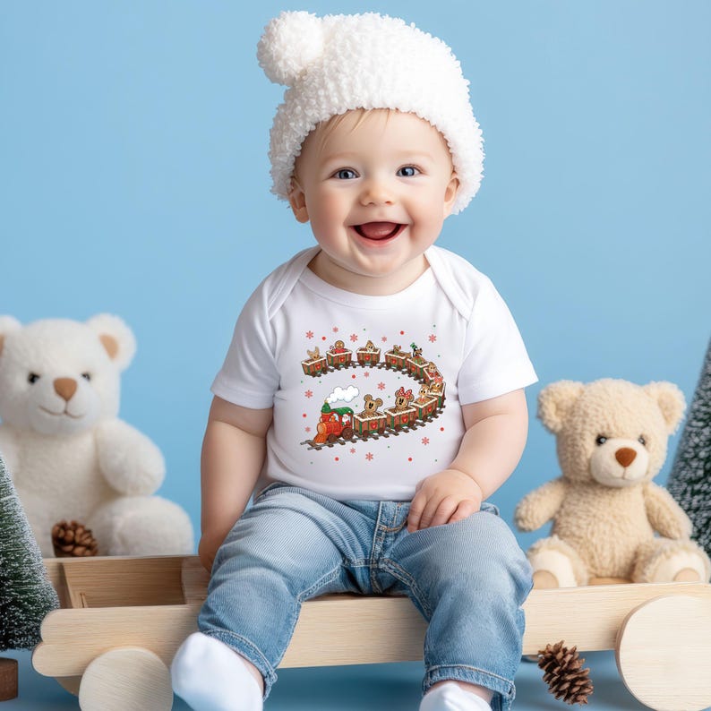 May include: A smiling baby wearing a white onesie with a Christmas train graphic, a white knit hat, and blue jeans. The baby is sitting on a wooden toy train with two teddy bears and a Christmas tree in the background.