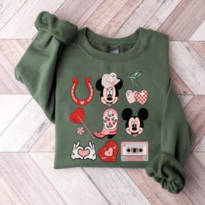 May include: A green sweatshirt featuring a graphic print of Mickey Mouse and Minnie Mouse in a country-themed design. The design includes a horseshoe, a cherry, a cowboy boot, a heart, a cassette tape, and a hand gesture of love.