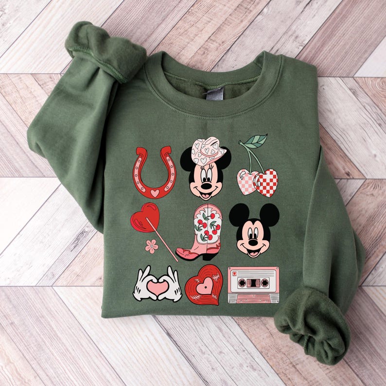May include: A green sweatshirt featuring a graphic print of Mickey Mouse and Minnie Mouse in a country-themed design. The design includes a horseshoe, a cherry, a cowboy boot, a heart, a cassette tape, and a hand gesture of love.