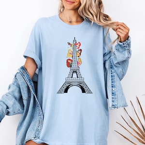 May include: Light blue t-shirt featuring a black Eiffel Tower design with cartoon princess illustrations. The graphic is centered on the shirt. The shirt has short sleeves and a crew neck.