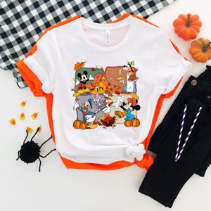 May include: White t-shirt with a colorful graphic featuring cartoon characters, pumpkins, and autumn leaves. The shirt has an orange trim and is paired with black pants. Decorative pumpkins and candy corn are also present.