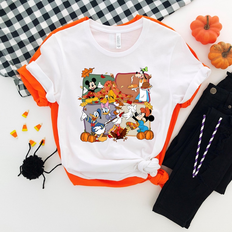 May include: White t-shirt with a colorful graphic featuring cartoon characters, pumpkins, and autumn leaves. The shirt has an orange trim and is paired with black pants. Decorative pumpkins and candy corn are also present.