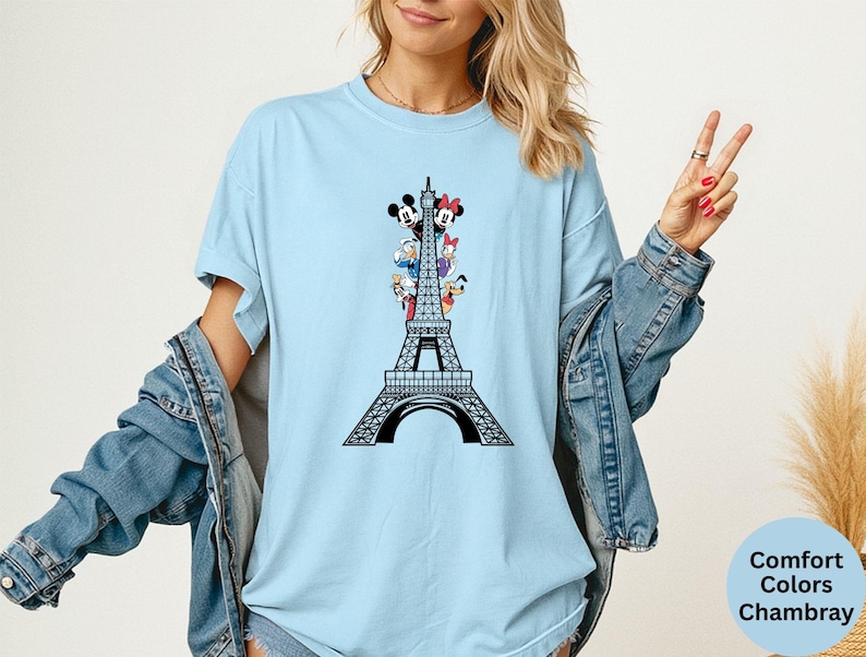 May include: Light blue t-shirt featuring a black Eiffel Tower design with Mickey Mouse, Minnie Mouse, Donald Duck, Daisy Duck, and Goofy. The shirt is made of a soft material and has a relaxed fit.