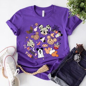 May include: Purple t-shirt with a spooky Mickey Mouse design. The design features Mickey Mouse and his friends dressed in Halloween costumes, surrounded by candy, pumpkins, and other Halloween-themed elements. The text "Spooky Vibes" is written on the shirt.