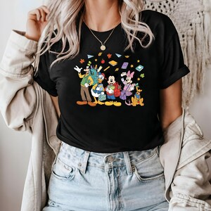 May include: Black t-shirt featuring a colorful graphic of Disney characters, including Goofy, Donald Duck, Mickey Mouse, Minnie Mouse, and Pluto, with books and autumn leaves. The characters are depicted with backpacks, suggesting a back-to-school theme.