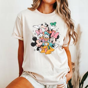 May include: A beige t-shirt with a graphic print of Mickey Mouse, Minnie Mouse, Donald Duck, Goofy, Pluto, and Daisy Duck. The characters are all smiling and have colorful accents.