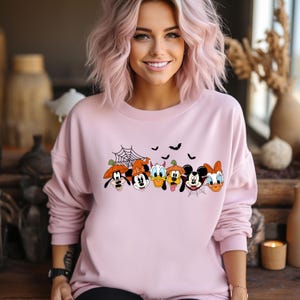 May include: A pink sweatshirt with a graphic print of Mickey Mouse, Minnie Mouse, Donald Duck, Goofy, Pluto, and Daisy Duck dressed in Halloween costumes. The characters are arranged in a row and are smiling. The sweatshirt has a crew neck and long sleeves.