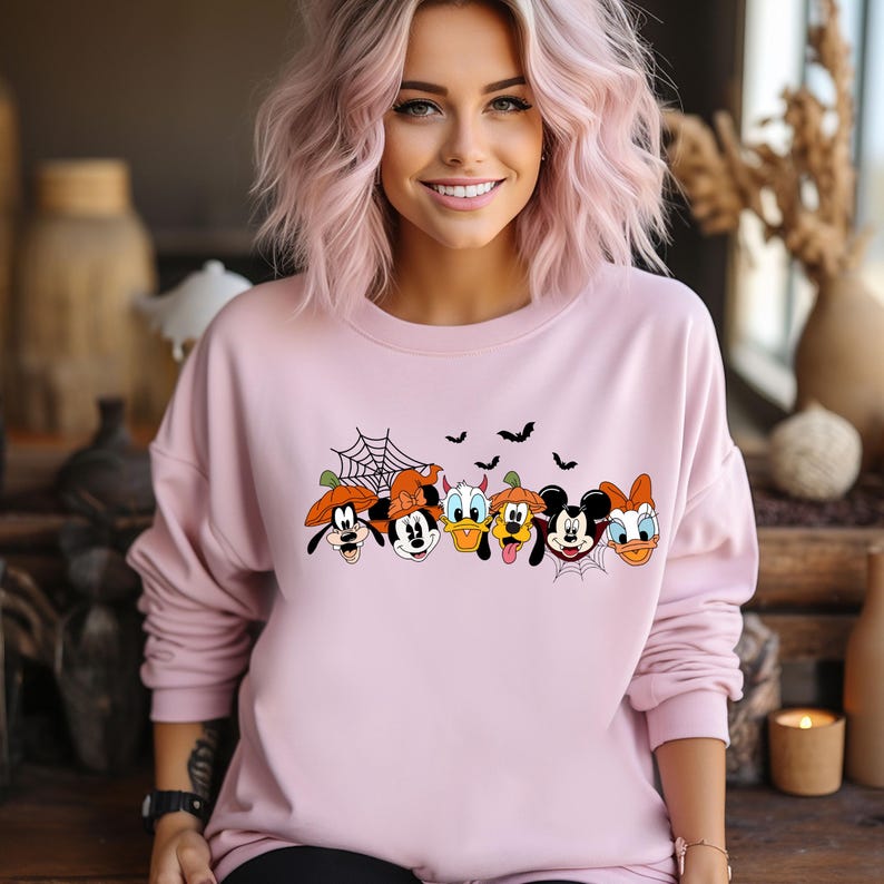 May include: A pink sweatshirt with a graphic print of Mickey Mouse, Minnie Mouse, Donald Duck, Goofy, Pluto, and Daisy Duck dressed in Halloween costumes. The characters are arranged in a row and are smiling. The sweatshirt has a crew neck and long sleeves.