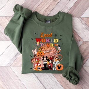 May include: Olive green sweatshirt featuring a colorful graphic of Disney characters in Halloween costumes. The graphic includes the text "Epcot World Tour 82" with bats and spiderwebs. The design includes Mickey and Minnie Mouse, Donald Duck, and Goofy.
