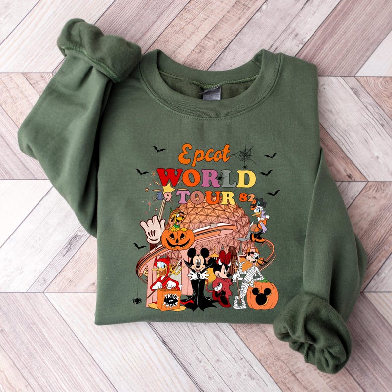 May include: Olive green sweatshirt featuring a colorful graphic of Disney characters in Halloween costumes. The graphic includes the text "Epcot World Tour 82" with bats and spiderwebs. The design includes Mickey and Minnie Mouse, Donald Duck, and Goofy.