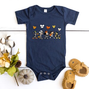 Op de afbeelding: Een donkerblauwe baby romper met een afbeelding van Mickey Mouse en zijn vrienden verkleed in Halloween kostuums. De personages houden ballonnen vast met Mickey Mouse oren. De romper heeft een drukknoop sluiting aan de onderkant.