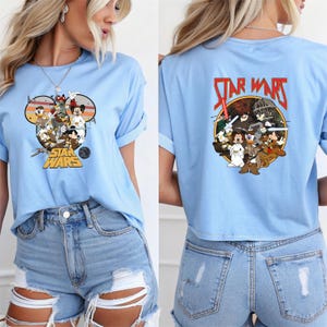 May include: Light blue t-shirt featuring a graphic of Disney characters in a Star Wars theme. The front design is within a Mickey Mouse head shape, and the back has a circular design with the Star Wars logo.