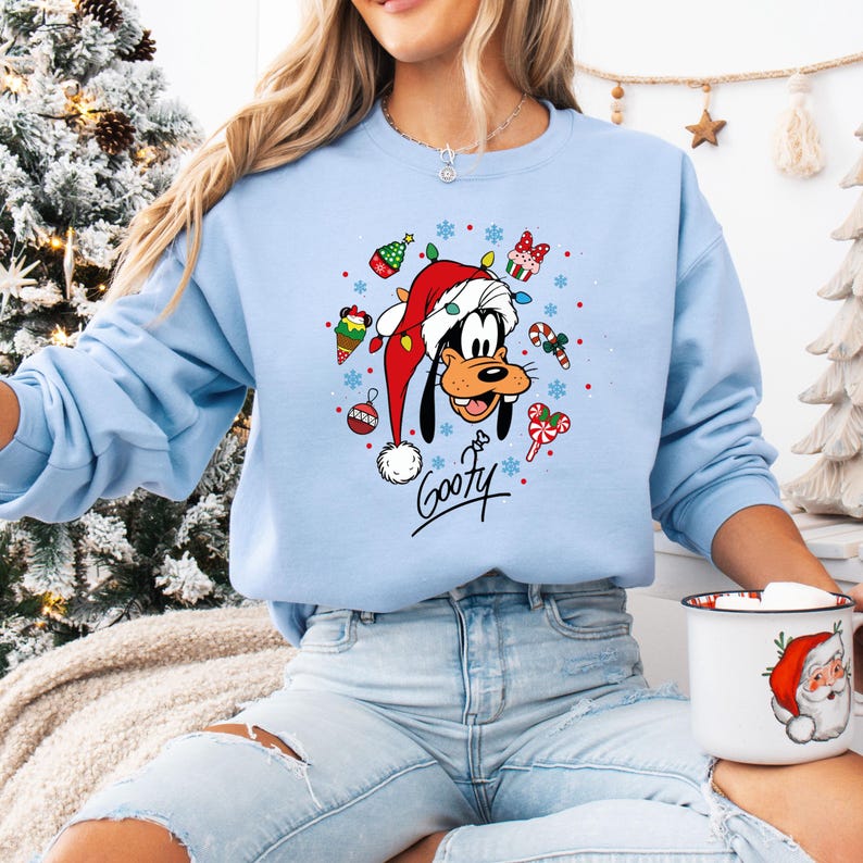 May include: Light blue sweatshirt featuring a graphic of Goofy wearing a Santa hat, surrounded by Christmas ornaments and candy canes. The word "Goofy" is written in a script font. A festive holiday-themed apparel item.