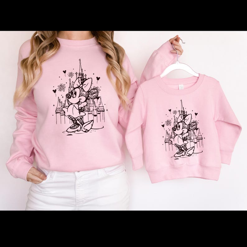 May include: Pink long-sleeved sweatshirts with a black and white illustration of Minnie Mouse in front of Cinderella's Castle.