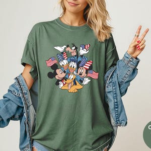 May include: Moss green t-shirt featuring a graphic of Mickey Mouse, Minnie Mouse, Donald Duck, Daisy Duck, Goofy, and Pluto holding American flags. The shirt has a crew neck and short sleeves. The text "Comfort Colors Moss" is in a circle on the bottom right.