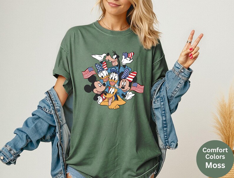 May include: Moss green t-shirt featuring a graphic of Mickey Mouse, Minnie Mouse, Donald Duck, Daisy Duck, Goofy, and Pluto holding American flags. The shirt has a crew neck and short sleeves. The text "Comfort Colors Moss" is in a circle on the bottom right.