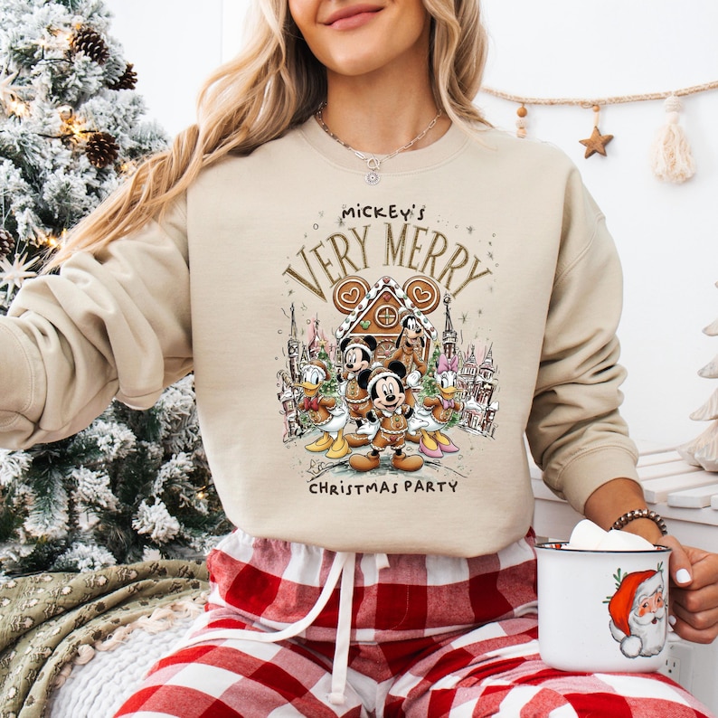 Puede incluir: Sudadera beige con un dise&ntilde;o festivo de Mickey Mouse y sus amigos. El dise&ntilde;o incluye una casa de jengibre y el texto "Mickey's Very Merry Christmas Party". La sudadera se combina con pantalones de pijama a cuadros rojos y blancos.