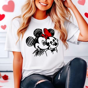 May include: White t-shirt featuring a black and white sketch of Mickey and Minnie Mouse embracing. Minnie Mouse is wearing a red bow. The design is a classic cartoon illustration.