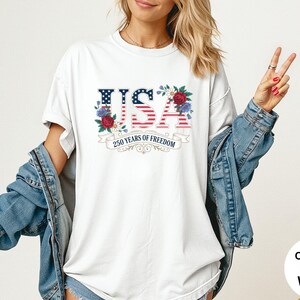 May include: White t-shirt with a patriotic USA design. The design features the word "USA" in red, white, and blue, with floral accents and the text "250 years of freedom". The shirt is part of the Comfort Colors collection.