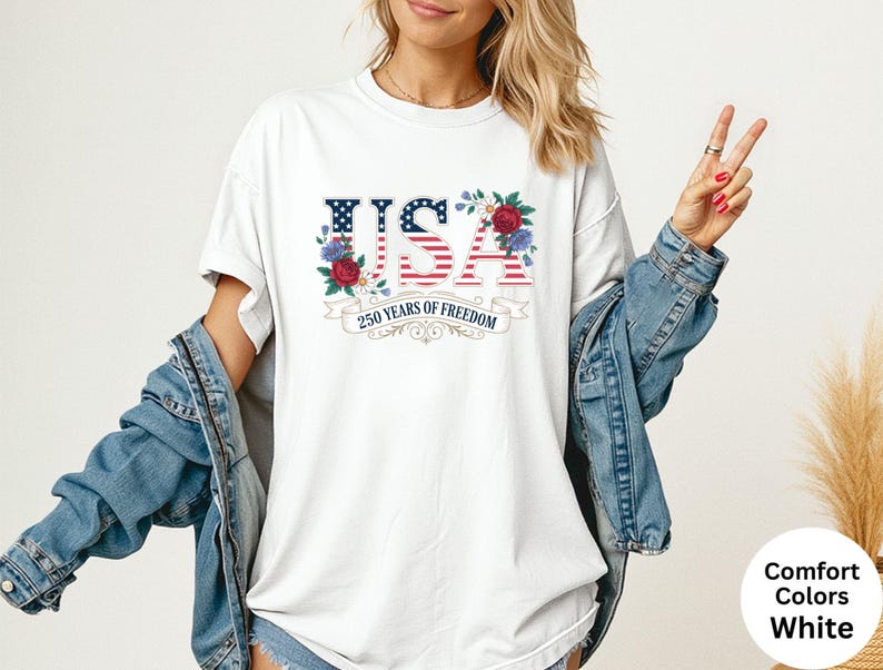 May include: White t-shirt with a patriotic USA design. The design features the word "USA" in red, white, and blue, with floral accents and the text "250 years of freedom". The shirt is part of the Comfort Colors collection.