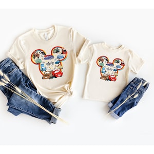 May include: Two cream colored t-shirts with a Mickey Mouse head design featuring a variety of cartoon cars in the ears. The larger shirt is for adults and the smaller shirt is for children.
