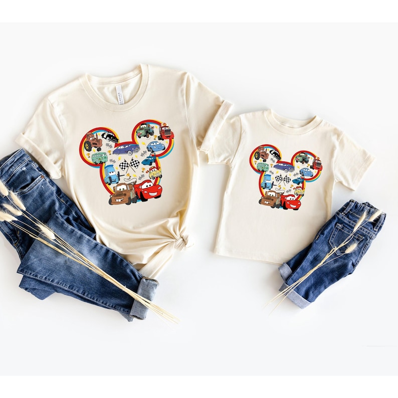 May include: Two cream colored t-shirts with a Mickey Mouse head design featuring a variety of cartoon cars in the ears. The larger shirt is for adults and the smaller shirt is for children.