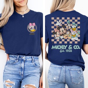 May include: A navy blue t-shirt with a checkered pattern on the back. The back of the shirt features a group of Disney characters including Mickey Mouse, Minnie Mouse, Donald Duck, Goofy, and Pluto. The text "MICKEY & CO. Est. 1928" is printed below the characters. The front of the shirt features a cartoon image of Daisy Duck with the text "ALICE" below.