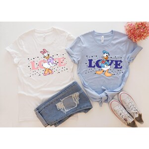 Op de afbeelding: Twee witte T-shirts met een blauwe en roze afbeelding van Donald Duck en Daisy Duck. De shirts hebben zwarte hartjes en het woord "LOVE" erop gedrukt in roze en blauw.