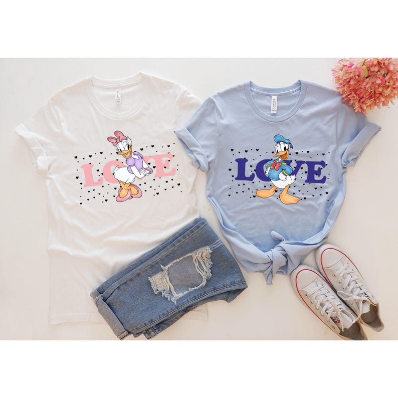 Op de afbeelding: Twee witte T-shirts met een blauwe en roze afbeelding van Donald Duck en Daisy Duck. De shirts hebben zwarte hartjes en het woord "LOVE" erop gedrukt in roze en blauw.