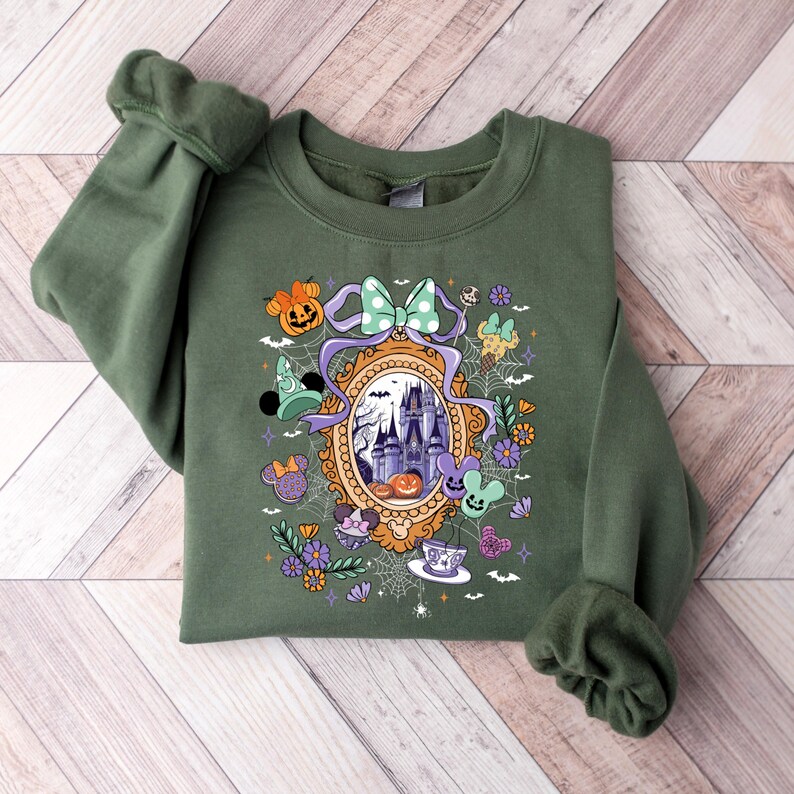 Puede incluir: Sudadera verde oliva con un dise&ntilde;o tem&aacute;tico de Halloween. El dise&ntilde;o incluye un castillo, calabazas, un lazo y varios elementos tem&aacute;ticos de personajes. La sudadera est&aacute; doblada sobre una superficie de madera.