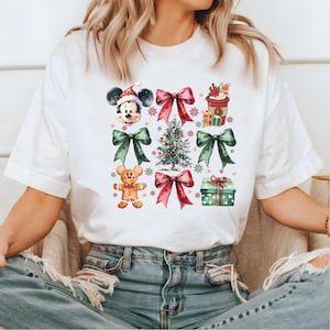 May include: White t-shirt with a Christmas-themed design featuring Mickey Mouse, a gingerbread man, a Christmas tree, bows, and a gift box.