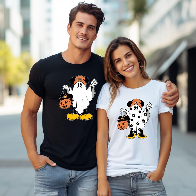 May include: Black and white t-shirts featuring a cartoon ghost character with Mickey Mouse ears, holding a jack-o'-lantern. The ghost wears a hat and yellow shoes. One shirt is black, the other white.