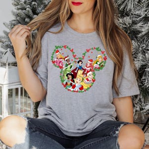 Pu&ograve; includere: Una maglietta grigia con un design festivo di principesse Disney con cappelli di Babbo Natale, disposte a forma di testa di Topolino. Le principesse sono circondate da una ghirlanda verde con accenti rossi, perfetta per le festivit&agrave;.