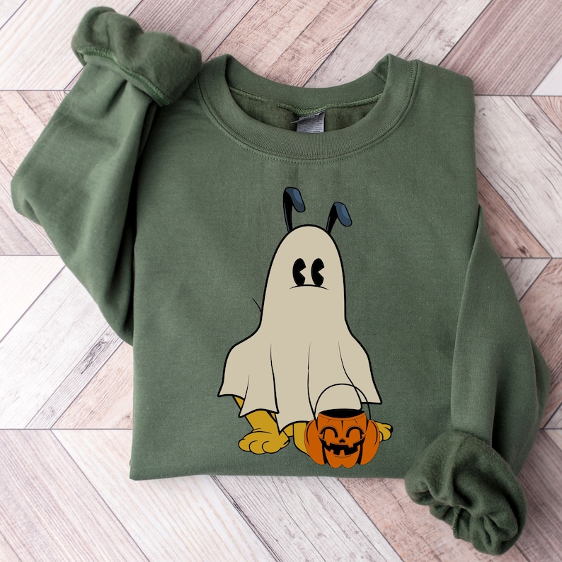 Pode incluir: Camisola verde-oliva com um design de c&atilde;o fantasma de desenho animado. O c&atilde;o est&aacute; usando uma fantasia de fantasma e segurando um balde de ab&oacute;bora. O design est&aacute; centrado na frente, perfeito para o Halloween.