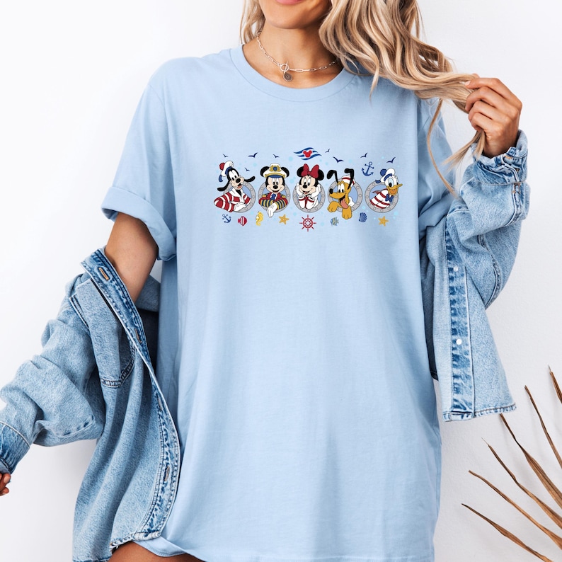 May include: Light blue t-shirt featuring a nautical-themed graphic of Disney characters including Goofy, Mickey, Minnie, Pluto, and Donald Duck. The characters are in circular frames with anchors, stars, and ship wheels.