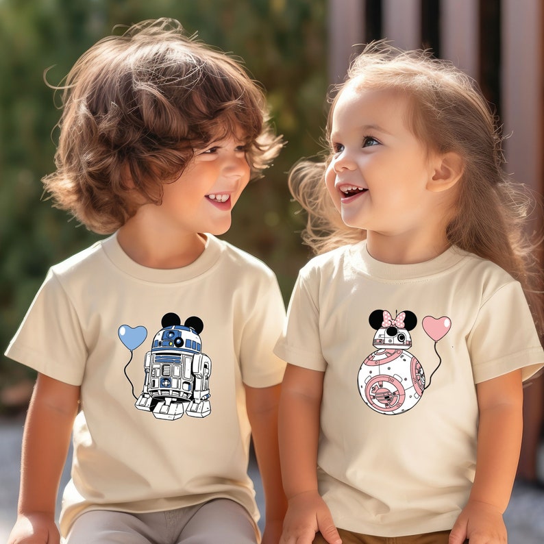 May include: Two children wearing beige t-shirts with Star Wars-themed graphics. One shirt features R2-D2 with Mickey Mouse ears and a blue heart balloon. The other shirt displays BB-8 with Mickey Mouse ears and a pink heart balloon.