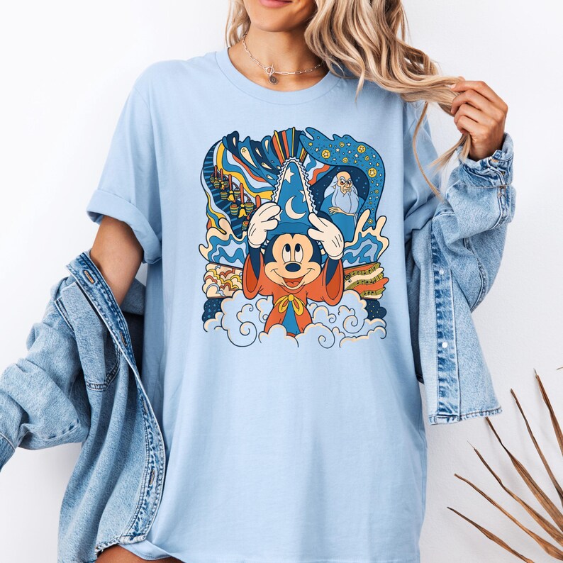 May include: Light blue t-shirt with a colorful graphic of Mickey Mouse as a sorcerer. The graphic features Mickey Mouse wearing a sorcerer's hat and robe, with a swirling background of stars, clouds, and other magical elements.