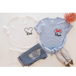 May include: Two t-shirts, one white and one light blue, with Disney-themed designs. The white shirt has a black Mickey Mouse head outline with the word "Dad." The blue shirt features a red polka-dot bow and the word "Mom." Blue jeans and white sneakers are also in the image.