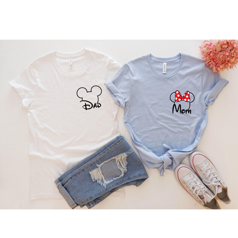 May include: Two t-shirts, one white and one light blue, with Disney-themed designs. The white shirt has a black Mickey Mouse head outline with the word "Dad." The blue shirt features a red polka-dot bow and the word "Mom." Blue jeans and white sneakers are also in the image.