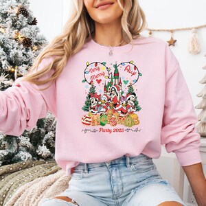 May include: A light pink sweatshirt featuring a colorful graphic of Disney characters in Christmas attire. The design includes the text "Very Merry" and "Christmas Party 2025". The sweatshirt has a crew neck and long sleeves.