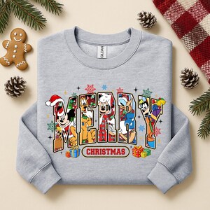 May include: A light gray sweatshirt with the word "MERRY" in colorful letters, each filled with a different Disney character in holiday attire. The word "CHRISTMAS" is below. Festive snowflakes and gift boxes surround the text.