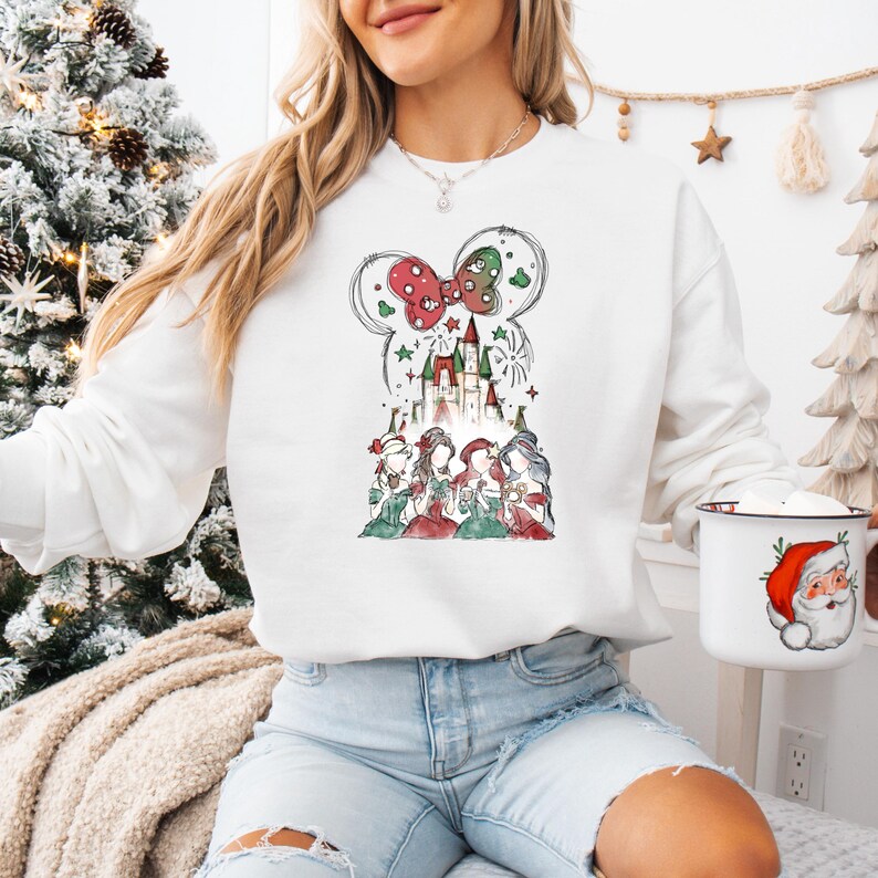 May include: White sweatshirt featuring a whimsical Christmas design. The graphic includes a castle, a bow, and several figures in festive attire. The design is in a watercolor style, with red, green, and gold accents. The sweatshirt is paired with denim shorts.