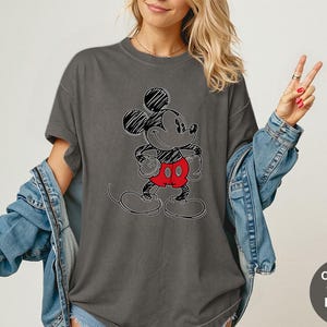 May include: Dark grey t-shirt featuring a sketched Mickey Mouse design with black outlines, red shorts, and black shoes. The shirt has short sleeves and a relaxed fit. The text "Comfort Colors Pepper" is in a circle on the bottom right.