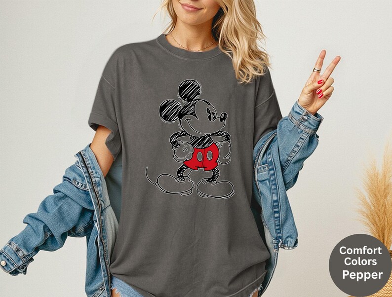 May include: Dark grey t-shirt featuring a sketched Mickey Mouse design with black outlines, red shorts, and black shoes. The shirt has short sleeves and a relaxed fit. The text "Comfort Colors Pepper" is in a circle on the bottom right.