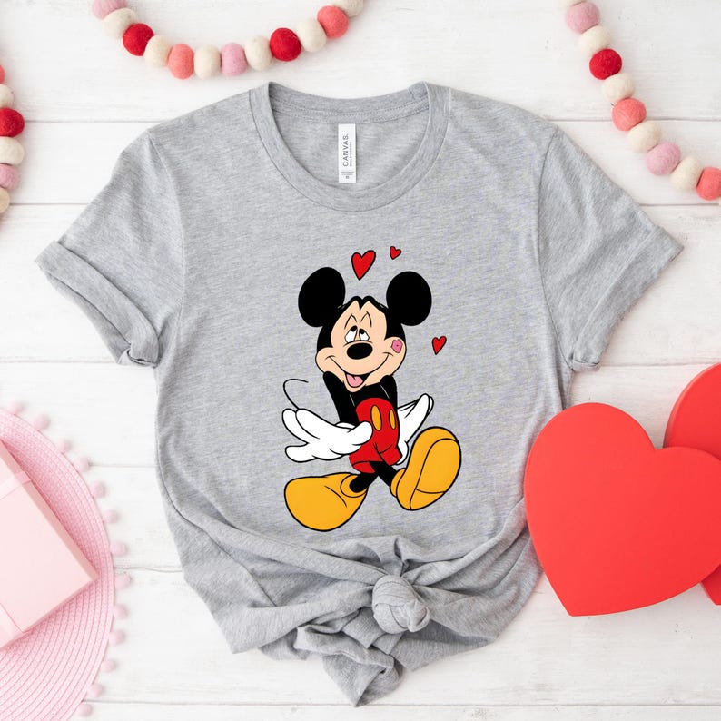 May include: A gray t-shirt featuring a cartoon Mickey Mouse design. Mickey is depicted with a red heart, surrounded by three small red hearts. The shirt has short sleeves and a knotted hem, perfect for casual wear.