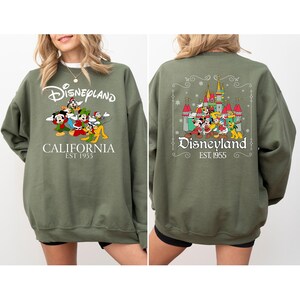 May include: Olive green Disneyland sweatshirt with front and back graphics. The front features the Disneyland logo, cartoon characters, and "CALIFORNIA EST 1955." The back displays the Disneyland castle, characters, and logo. A cozy, long-sleeved top.