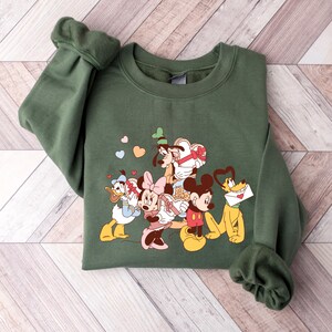 May include: A green sweatshirt with a graphic print of Mickey Mouse, Minnie Mouse, Donald Duck, Goofy, and Pluto. The characters are holding hearts and gifts.